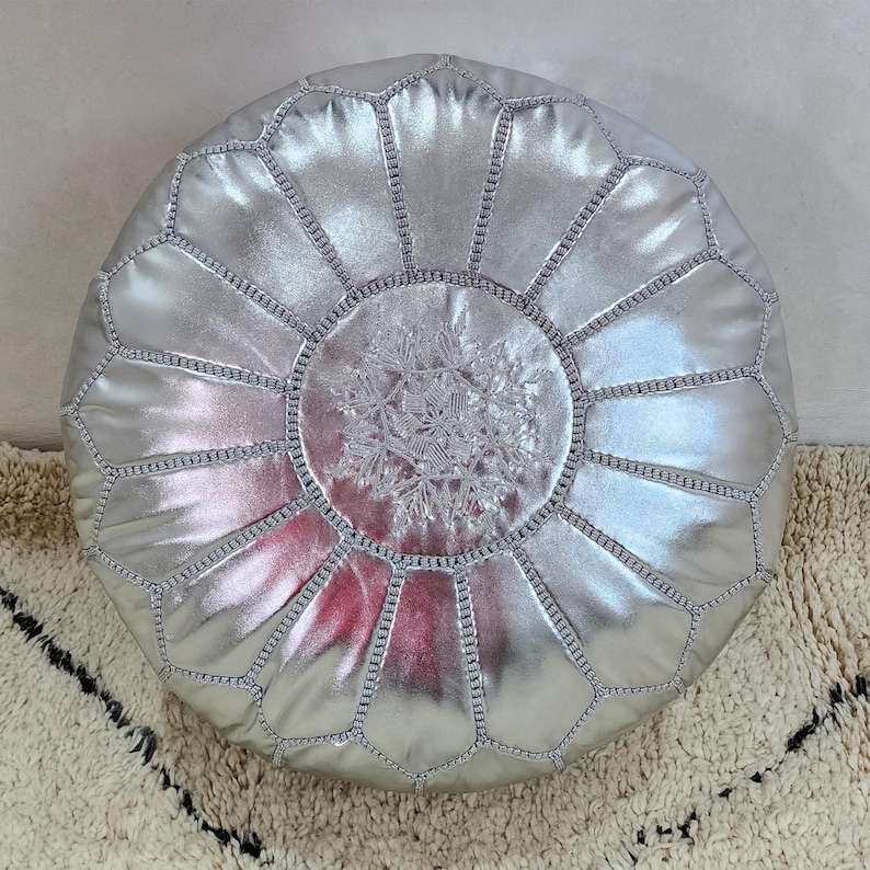 May include: A round, silver pouf with a metallic sheen. The top features a detailed, embroidered snowflake design in a lighter silver thread. The pouf is constructed with geometric panels and visible stitching.