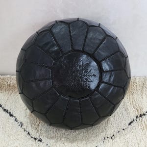 May include: A round, black leather pouf with a geometric pattern. The pouf is made of stitched panels and features a central embossed design. The pouf is placed on a cream-colored rug with a black geometric pattern.