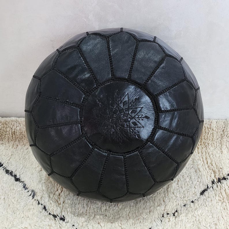 May include: A round, black leather pouf with a geometric pattern. The pouf is made of stitched panels and features a central embossed design. The pouf is placed on a cream-colored rug with a black geometric pattern.