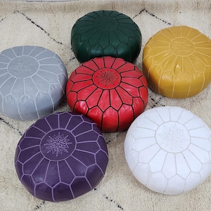 May include: Six round Moroccan leather poufs in various colors: gray, green, red, yellow, white, and purple. Each pouf features a geometric design and decorative stitching. The poufs are arranged on a patterned rug, showcasing their vibrant colors and textures.