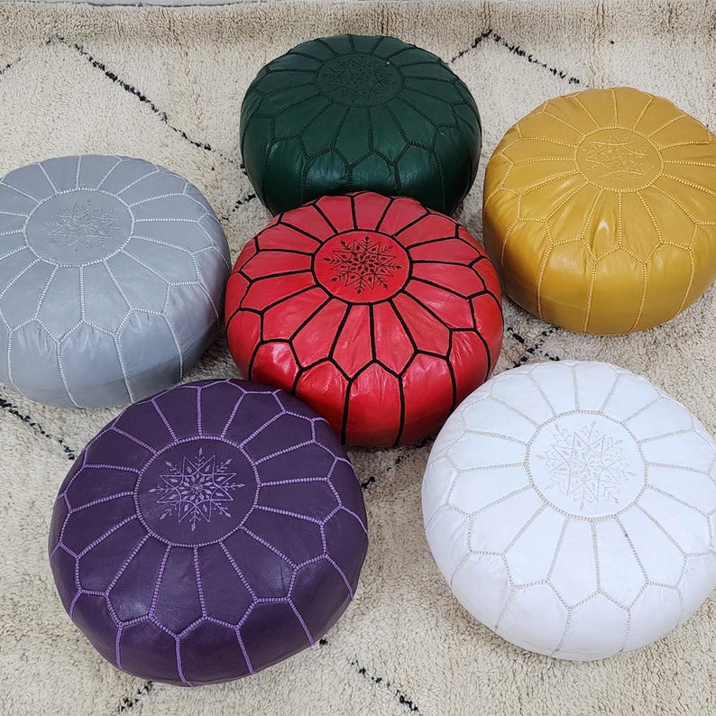 May include: Six round Moroccan leather poufs in various colors: gray, green, red, yellow, white, and purple. Each pouf features a geometric design and decorative stitching. The poufs are arranged on a patterned rug, showcasing their vibrant colors and textures.