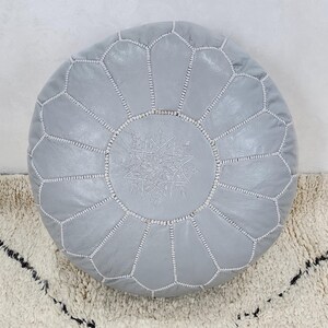 May include: A round, light gray leather pouf with a geometric pattern and white stitching. The center features a decorative embroidered design. The pouf is placed on a textured, off-white rug with black accents.