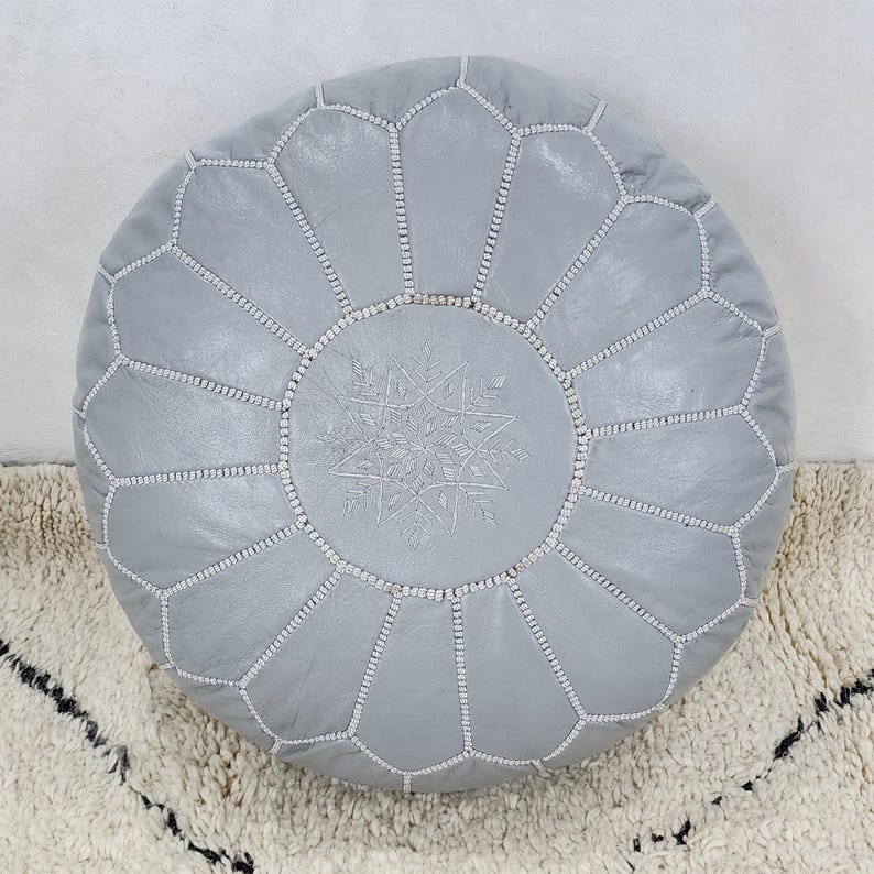 May include: A round, light gray leather pouf with a geometric pattern and white stitching. The center features a decorative embroidered design. The pouf is placed on a textured, off-white rug with black accents.