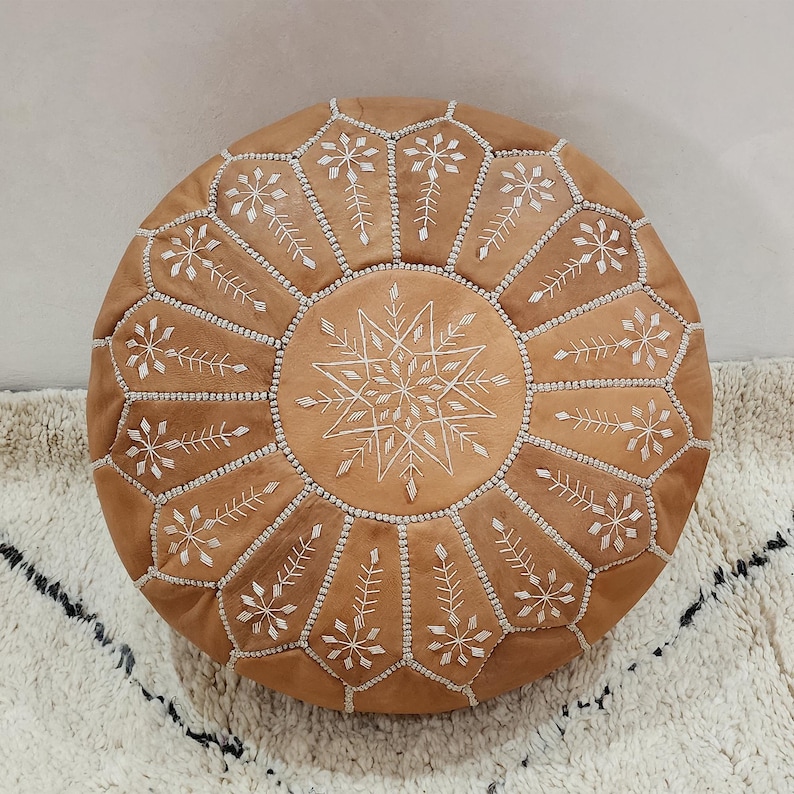 May include: Round, tan leather pouf with white embroidered geometric patterns. The pouf features a central starburst design and is surrounded by floral and leaf motifs. The stitching is white, and the pouf is placed on a white rug with black accents.