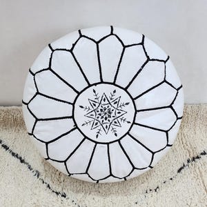 May include: White round Moroccan leather pouf with black geometric stitching. The center features a black starburst design. This decorative pouf can be used as a footstool or extra seating. The pouf is approximately 20 inches in diameter.