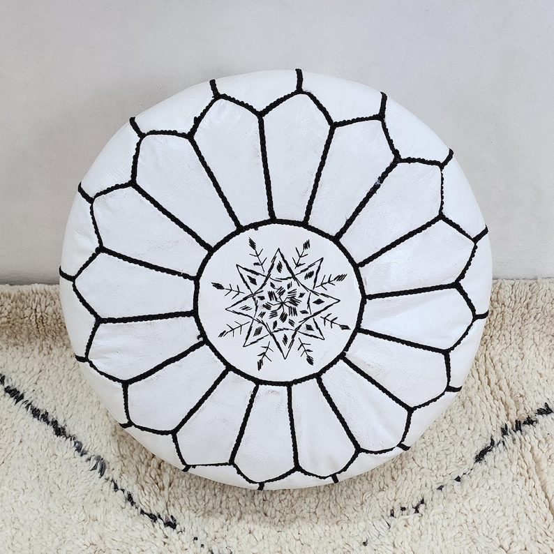 May include: White round Moroccan leather pouf with black geometric stitching. The center features a black starburst design. This decorative pouf can be used as a footstool or extra seating. The pouf is approximately 20 inches in diameter.