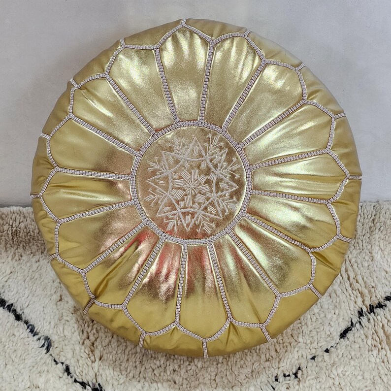 May include: A round, gold-colored pouf with a metallic sheen. The pouf features a geometric design with stitched seams and a central embroidered snowflake pattern. The pouf is placed on a cream-colored rug with a black geometric pattern.