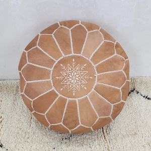 May include: A round, tan leather pouf with white stitching and a geometric pattern. The center features a white starburst design. This decorative floor cushion adds a touch of bohemian style to any living space.