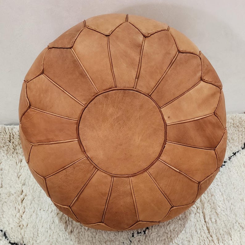 May include: Round, light brown leather ottoman with a geometric design. The ottoman is made of stitched leather panels, creating a flower-like pattern. It is placed on a white rug with black accents. This decorative piece adds a touch of bohemian style to any living space.