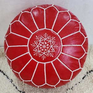 May include: A round, red leather pouf with white stitching and a white snowflake-like design in the center. The pouf is a decorative piece, likely for seating or as a footrest. The design is geometric and the color is a vibrant red.