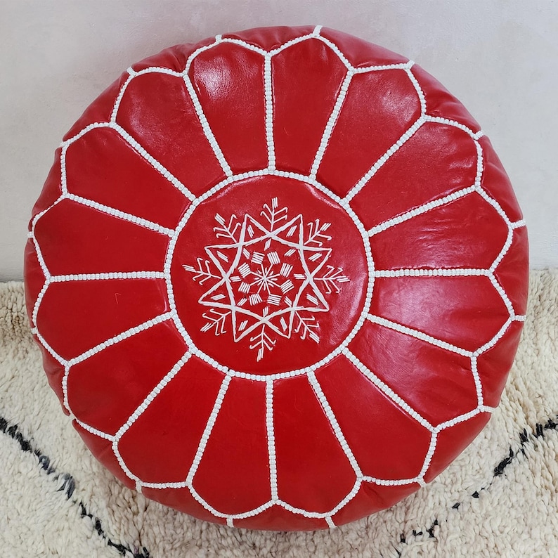May include: A round, red leather pouf with white stitching and a white snowflake-like design in the center. The pouf is a decorative piece, likely for seating or as a footrest. The design is geometric and the color is a vibrant red.