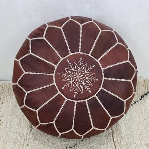 May include: Round, brown leather pouf with white stitching and a snowflake-like design in the center. The pouf is placed on a white rug with black accents. This decorative piece adds a touch of bohemian style to any living space.