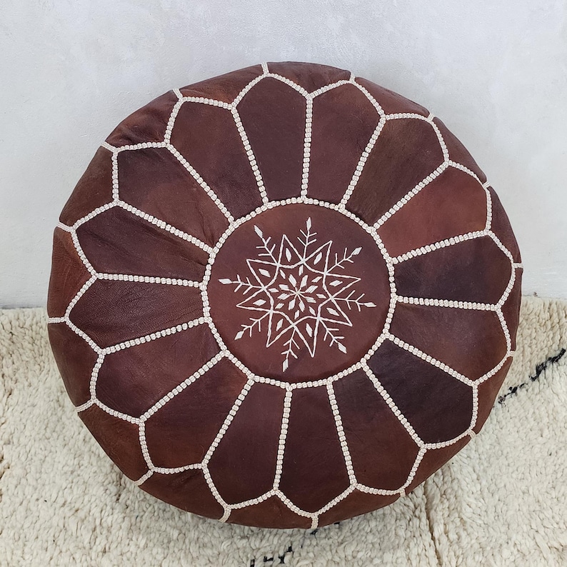 May include: Round, brown leather pouf with white stitching and a snowflake-like design in the center. The pouf is placed on a white rug with black accents. This decorative piece adds a touch of bohemian style to any living space.