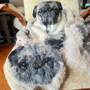 May include: A pug dog is nestled in a chair, covered by a patterned blanket. A pug-shaped pillow rests in front. The blanket has a mix of pink, grey, and cream tones. The scene evokes comfort and warmth, with the dog as the focal point.