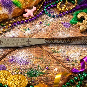 May include: A silver knife with a decorative handle and the text "KING CAKE'S KNIFE STAYS IN THE BOX" is surrounded by Mardi Gras beads, coins, and a golden fleur-de-lis on a wooden surface. The scene is festive and colorful.