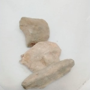 May include: Three irregularly shaped, light beige stones are arranged on a white surface. The stones have a rough, textured appearance, with variations in color from light tan to a slightly darker shade.