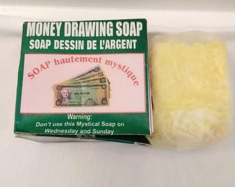 May include: A box of "Money Drawing Soap" with text in English and French, and a bar of yellow soap. The box has a green border and a pink center with an image of money. The warning states not to use the soap on Wednesday and Sunday.