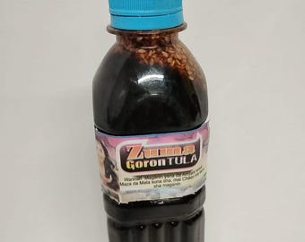 May include: A clear plastic bottle filled with a dark liquid, topped with a blue cap. The bottle has a label with the text "Zuma Goron Tula" and additional text in a foreign language. The liquid appears to contain seeds.