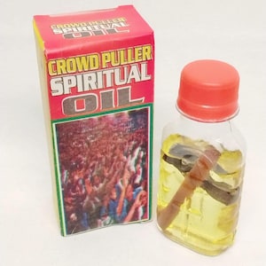 May include: A small, clear glass bottle with a red cap, filled with yellow liquid and a brown stick. Beside it is a red, yellow, and green box with the words "CROWD PULLER SPIRITUAL OIL" printed on it. The box features an image of a crowd.