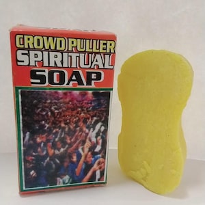 May include: A yellow, kidney-shaped bar of soap next to a box. The box is red with the words "CROWD PULLER SPIRITUAL SOAP" in white and yellow. The box has a picture of a crowd of people.