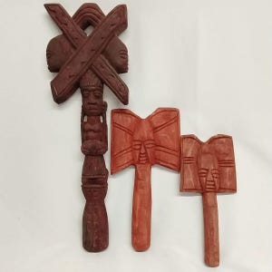 May include: Three carved wooden sculptures in shades of red and brown. The largest is a totem-like figure with a cross-shaped top and a carved face. Two smaller sculptures have axe-like shapes with carved faces.