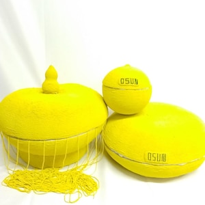 May include: Three bright yellow decorative objects. The largest is a round bowl with a lid and a tassel. The middle object is a small sphere. The bottom object is a flat, round shape. All objects have the word "OSUN" printed on them.