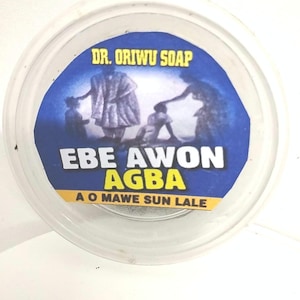 Peut inclure: Un récipient rond en plastique transparent avec une étiquette bleue et jaune. L'étiquette porte le texte "DR. ORIWU SOAP" et "EBE AWON AGBA" avec une illustration de personnages. Le récipient est sur une surface blanche.