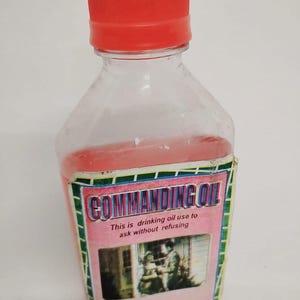 May include: A clear plastic bottle with a red cap and a pink label that reads "COMMANDING OIL This is drinking oil use to ask without refusing Products of Dr. Robert".