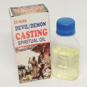 May include: A clear glass bottle with a blue cap next to a white box. The box has text that reads "DR WURA DEVIL/DEMON CASTING SPIRITUAL OIL" in red and blue. The bottle contains a yellow liquid.