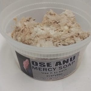 May include: A clear plastic container filled with a light brown, textured soap. The container has a label with the text "OSE ANU MERCY SOAP" and the words "Use it to bath everyday".