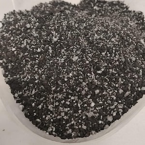 May include: Close-up of a pile of dark gray and white granular material. The material appears to be a mix of small, irregularly shaped pieces, with a speckled appearance. The texture is coarse and uneven.