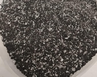 May include: Close-up of a pile of dark gray and white granular material. The material appears to be a mix of small, irregularly shaped pieces, with a speckled appearance. The texture is coarse and uneven.