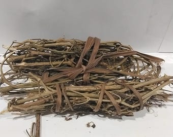 May include: A bundle of dried, thin, brown twigs tied together with a woven, light brown material. The twigs are of varying lengths and thicknesses, creating a textured appearance. The bundle is resting on a white surface.