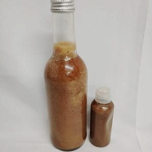 Op de afbeelding: Twee heldere glazen flessen, een lange en een kleine, gevuld met een bruine vloeistof. De lange fles heeft een zilveren dop en de kleine fles heeft een witte dop. De vloeistof heeft een korrelige textuur.