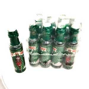 Può includere: Un gruppo di piccole bottiglie di vetro verde con tappi bianchi, avvolte in plastica trasparente. Ogni bottiglia ha un'etichetta con testo e un'illustrazione di una persona. Le bottiglie sono disposte in gruppo, con una bottiglia separata.