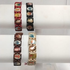 May include: Four religious bracelets with rectangular wooden beads. Two bracelets are red and black, and two are brown and light wood. Each bead features a religious image. The bracelets are displayed on a white surface.