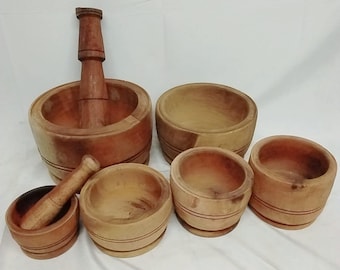 May include: A collection of wooden kitchen tools, including a mortar and pestle set and several small bowls. The wood has a warm, natural tone. The mortar and pestle are larger than the bowls, which vary in size.