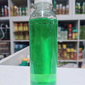 May include: A clear plastic bottle with a white cap, filled with a vibrant green liquid. The bottle is cylindrical and sits on a white surface. The background shows shelves with various bottles.