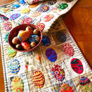 May include: A quilted table runner featuring a patchwork design of various fabric squares with printed text and colourful oval-shaped egg appliqués. A dark red ceramic bowl filled with several painted wooden eggs sits on the runner.