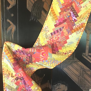Puede incluir: Un camino de mesa de tela largo y rectangular con un vibrante diseño de patchwork de varios textiles estampados en amarillo, naranja, rojo y rosa. El camino está rematado con un borde fino a rayas amarillas y naranjas en todos los lados.