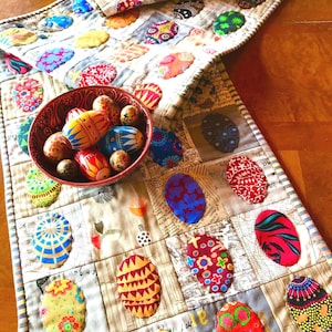 May include: A quilted table runner featuring a patchwork design of various colourful egg-shaped fabric appliqués in diverse patterns, paired with a dark red bowl filled with several small, hand-painted wooden eggs in vibrant colours.