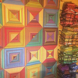 May include: A large rectangular quilt featuring a repeating geometric pattern of concentric squares in a vibrant spectrum of colors including red, orange, yellow, green, blue, and purple. Beside the quilt are four stacked wire baskets filled with fabric.