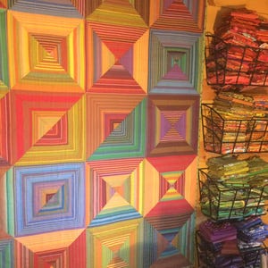 May include: A quilt featuring a geometric pattern of concentric squares in a vibrant spectrum of colors including red, orange, yellow, green, blue, and purple. Beside the quilt are four stacked wire baskets filled with folded fabric scraps.