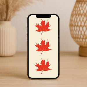May include: A smartphone displays three red maple leaf illustrations on a cream background. The phone is black and sits on a light brown wooden surface. A white vase with dried plants and a woven basket are in the background.