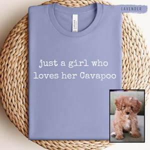 Op de afbeelding: Een lavendelkleurig T-shirt met de witte tekst "just a girl who loves her Cavapoo". Een kleine afbeelding van een Cavapoo-hond staat in de rechter benedenhoek. Het shirt is op een geweven oppervlak gevouwen.