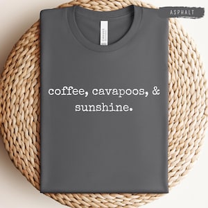 Peut inclure: T-shirt gris foncé avec l'inscription blanche "coffee, cavapoos, & sunshine". Le t-shirt a un col rond et est fait d'une matière douce. Le t-shirt est plié sur une surface tissée.