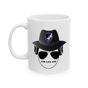 May include: White ceramic mug featuring a cartoon design. The design includes a black fedora hat with a shield emblem, sunglasses, headphones, and a stylized face. The mug has a white handle and a white interior.