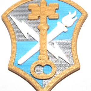 May include: A shield-shaped emblem featuring a gold border and a divided background of light blue and silver. Centered is a gold key overlaid on two crossed white lightning bolts and a white torch with a flame at the top.