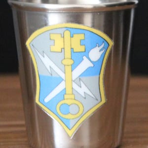 May include: A stainless steel shot glass with a yellow, blue, and gray military unit insignia featuring a key, lightning bolt, and torch.