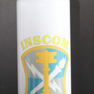 May include: White water bottle with a gold-colored bullet-shaped lid. The bottle features the text "INSCOM VETERAN" in yellow, along with a shield emblem with a key and lightning bolts.